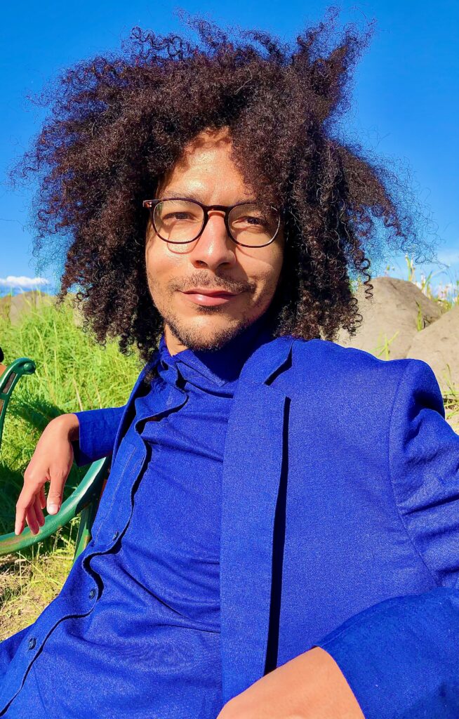 An man with an Afro seated on a bench on a clear summer day. He smiles and wears a blue suit.